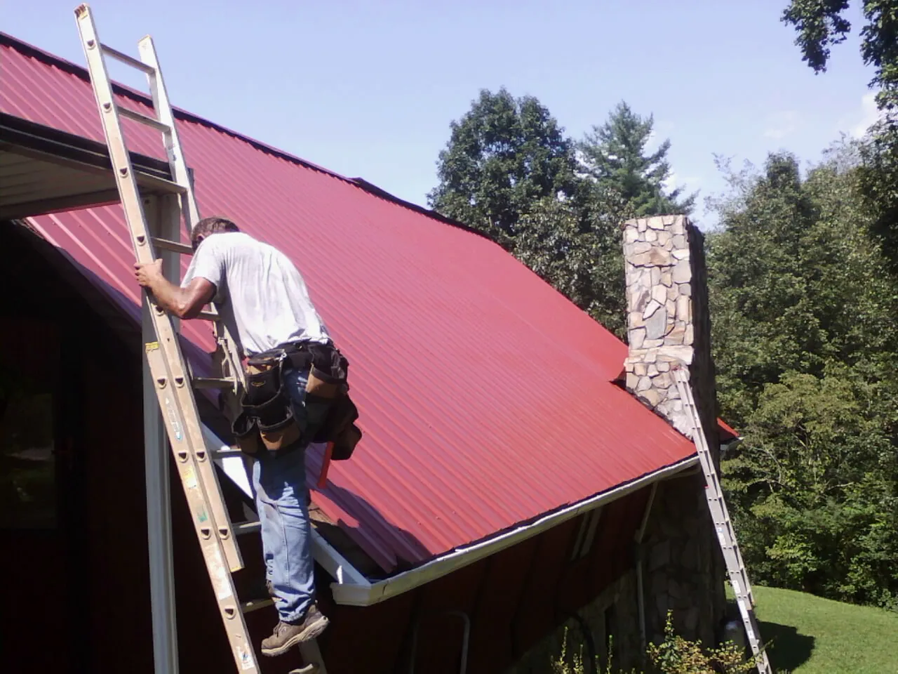 Roofer on a ladder beside a red metal roof and stone chimney — Soffit & Fascia Repair by Roofing Brothers in Climax